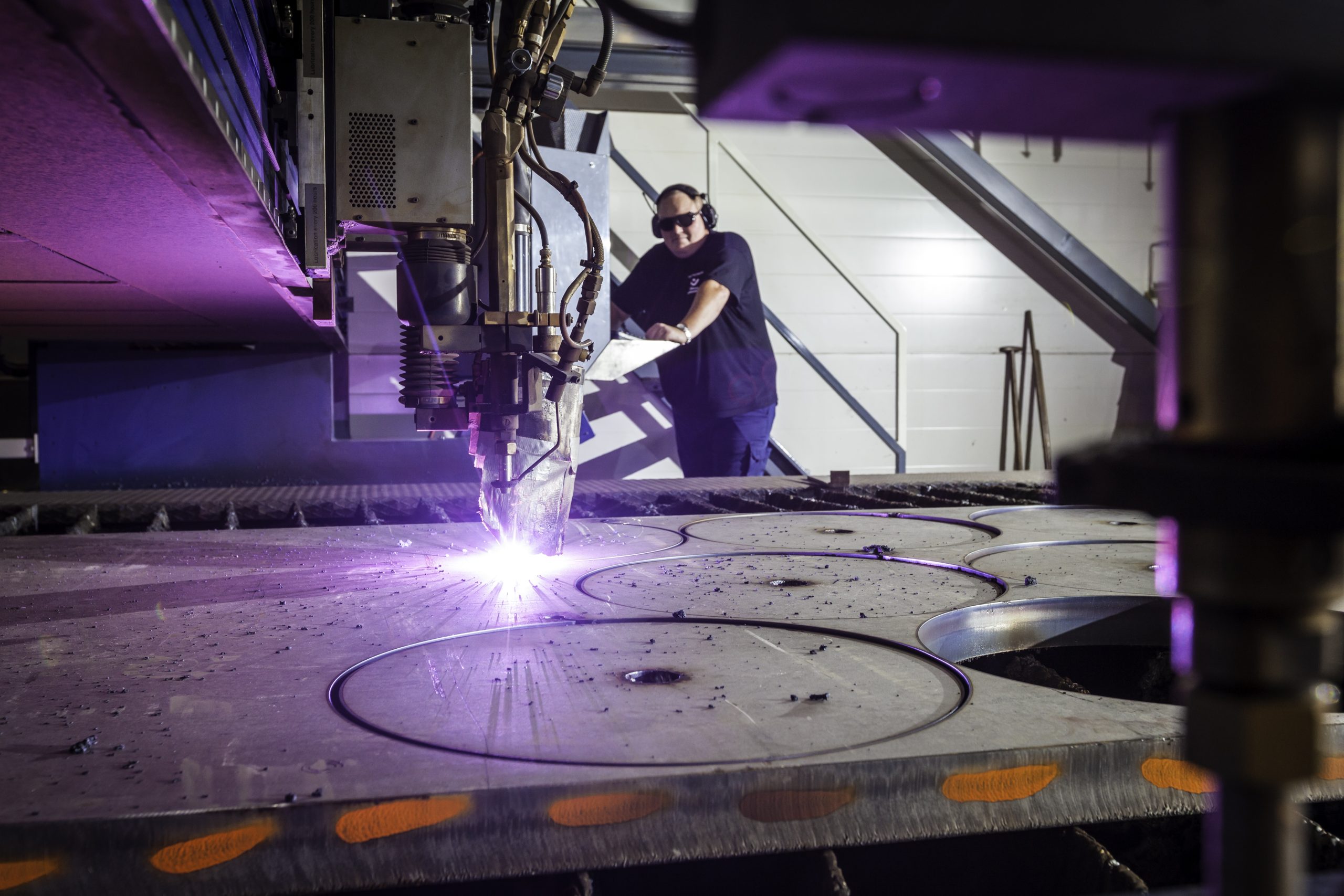 Plasmaskæring udført af certificeret i medarbejder, der står i baggrunden og overser processen - Viser laserskæreren i gang, med blå laserflamme imens der skæres runde elementer ud i metal